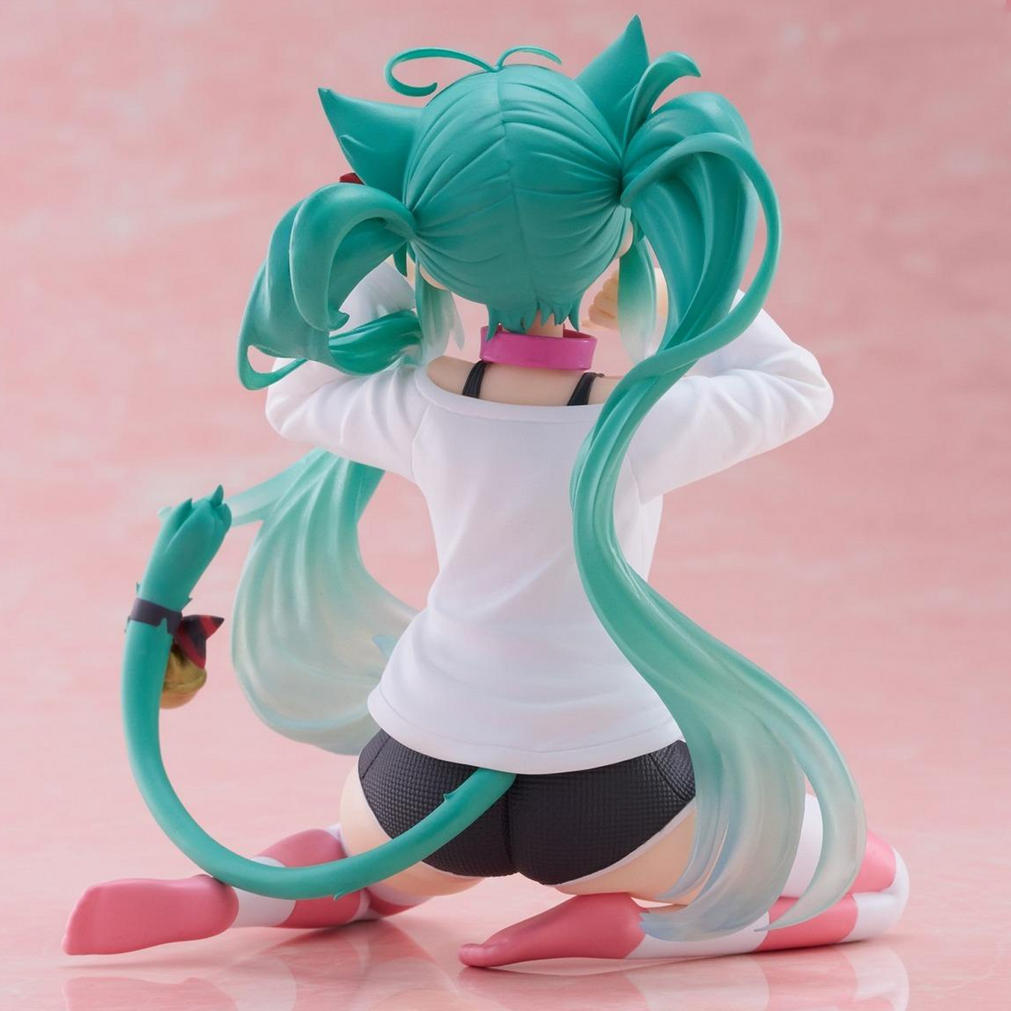 A turquoise-haired anime-style figurine shown from the back, kneeling on a pink surface against a pink background. The figure wears a white top, dark gray shorts, and pink-and-white shoes, with a long curved tail and cat-ear hair details | Hatsune Miku Desktop Cute Nekomimi T-Shirt ver. Figure | Happy Piranha