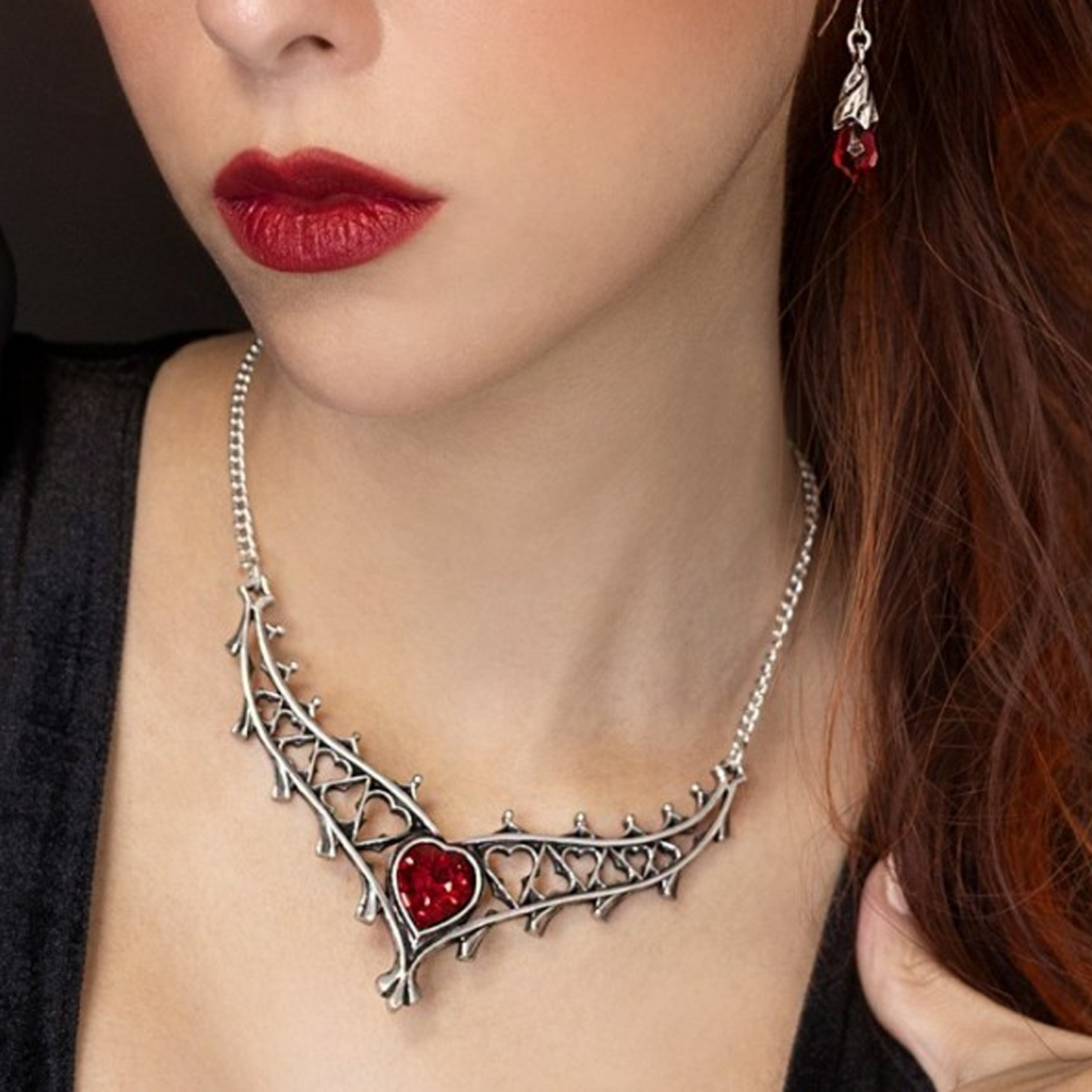 Close-up of a woman with red lipstick wearing a silver gothic arch design necklace with red heart-shaped stone in the centre and matching earrings | Happy Piranha