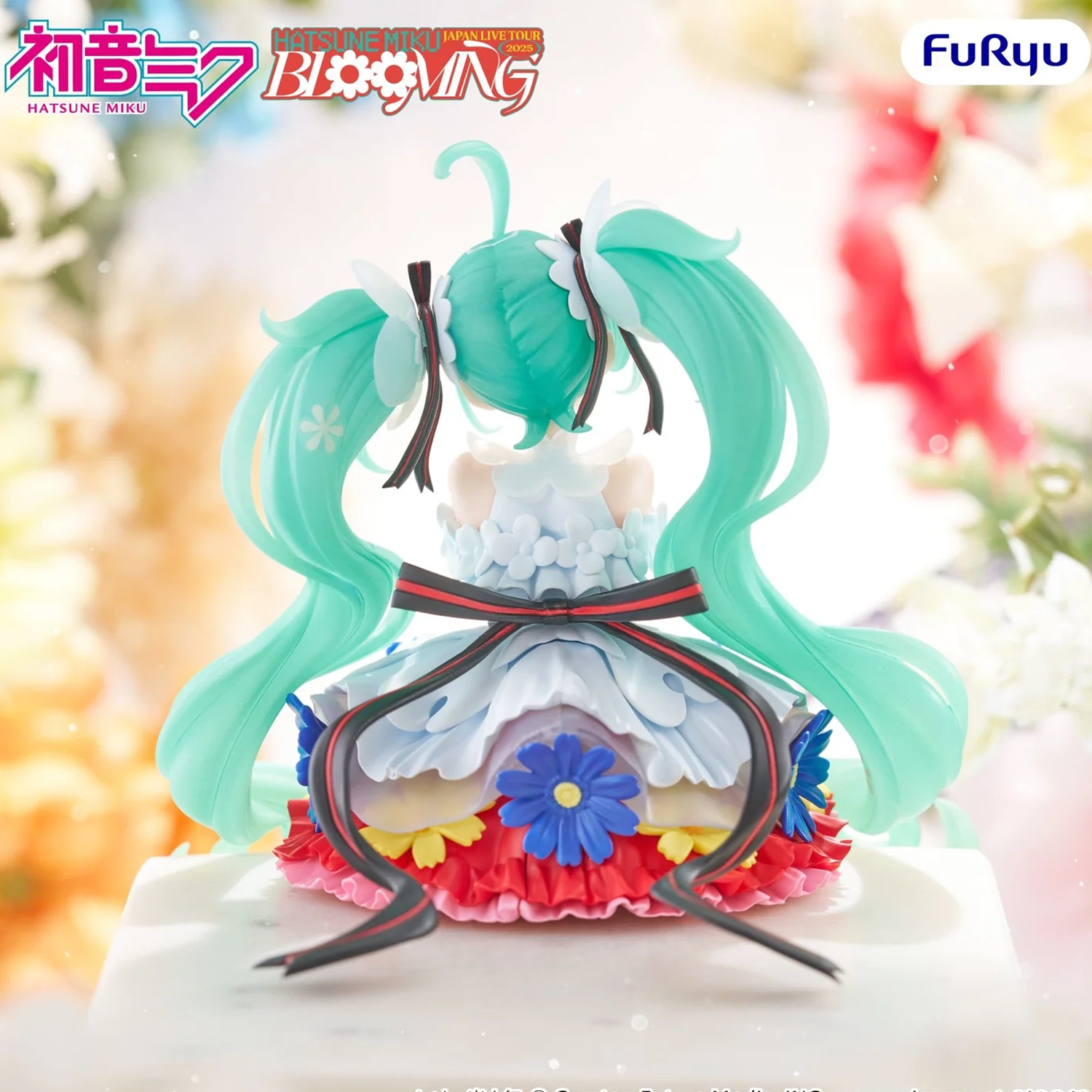 A Hatsune Miku 'Blooming' figure shown from the back, featuring signature mint green twin-tails and a layered white, blue, yellow, and red dress, set against a soft, blurred floral background. The 'Hatsune Miku Bloomin'' logo is visible in the top-left corner, and the 'FuRyu' brand logo is in the top-right | Hatsune Miku Japan Live Tour 2025 Blooming Noodle Stopper Figure | Happy Piranha