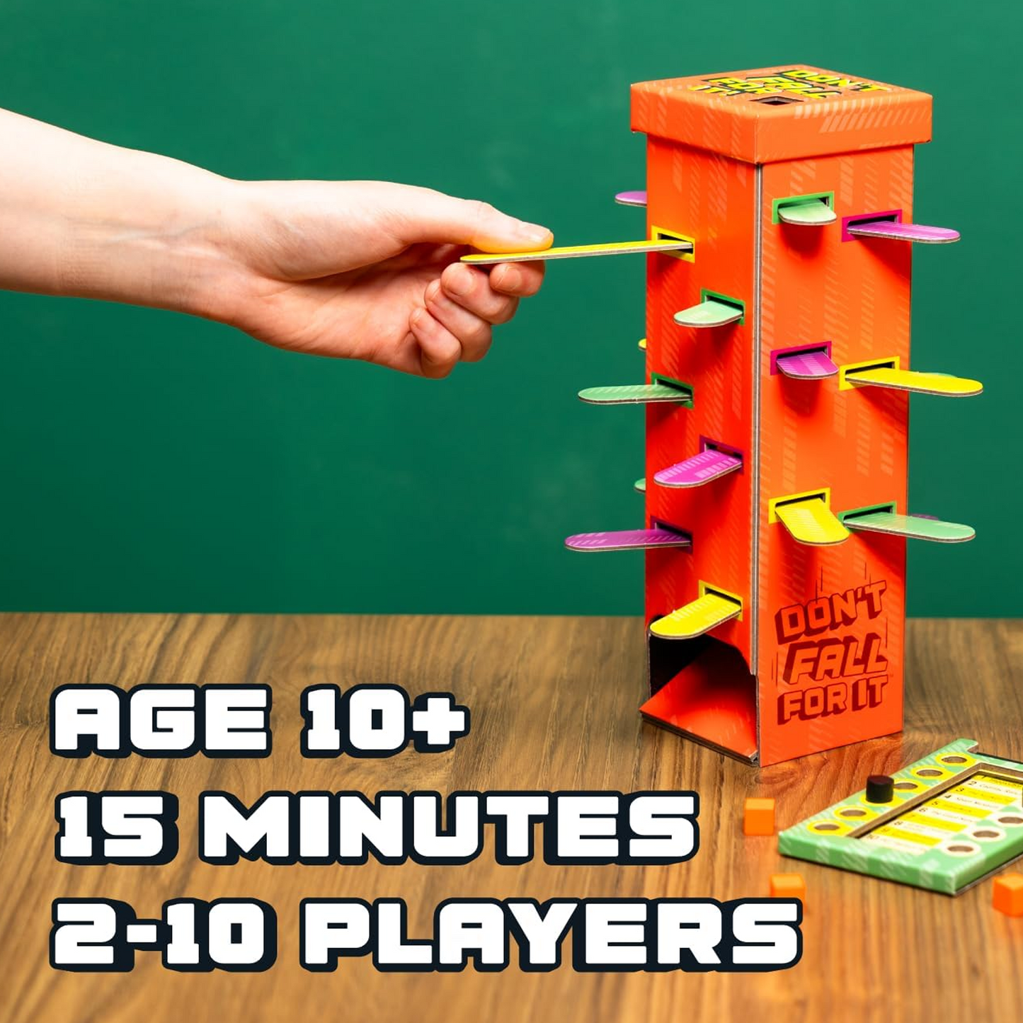 A Hand Pulling a Piece out of the Don't Fall For it Board Game Tower on a Wooden Table with as Green Wall Behind | Happy Piranha