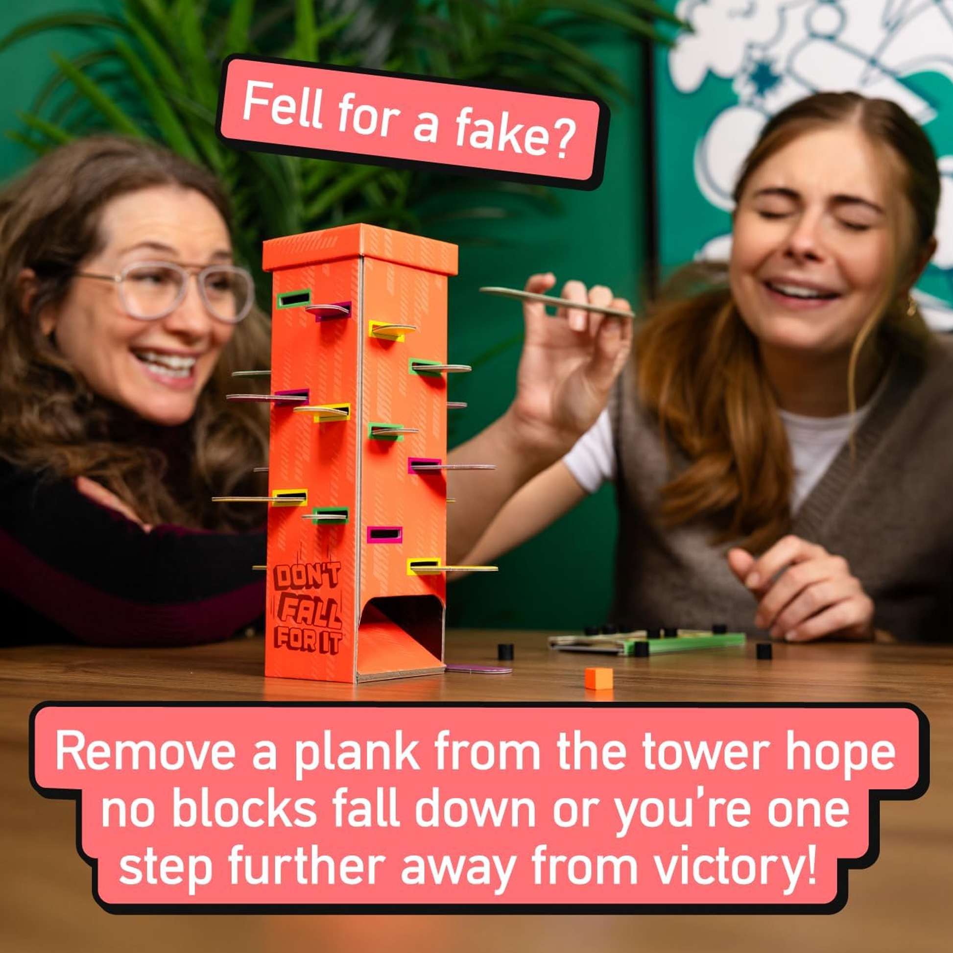 Two Women Laughing and Playing Don't Fall For It! Board Game on Wooden Table with a Green Wall and Plant in the Background | Happy Piranha