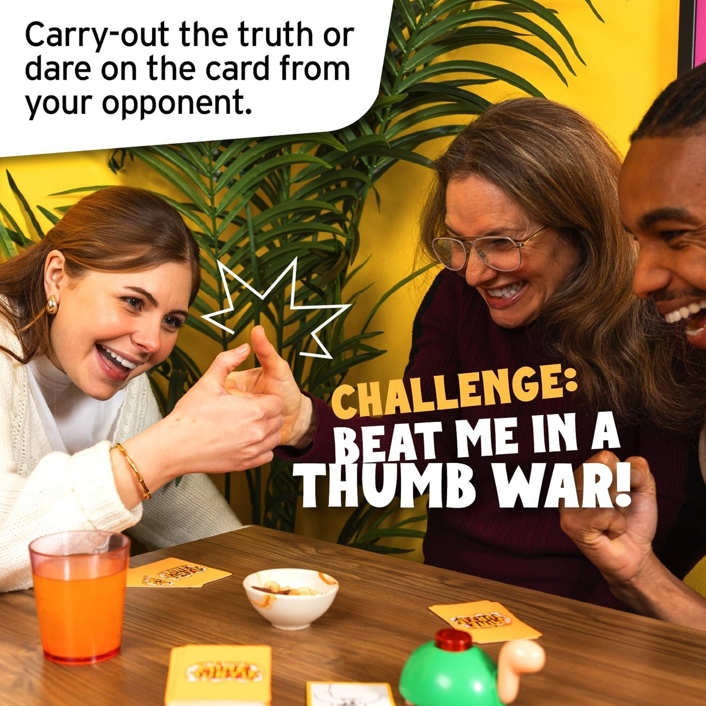 2 Women and a Man Laughing and Playing Don't Press That Mine Turtle While Having a Thumb War on a Wooden Table with a Plant and Yellow Wall in the Background  | Happy Piranha