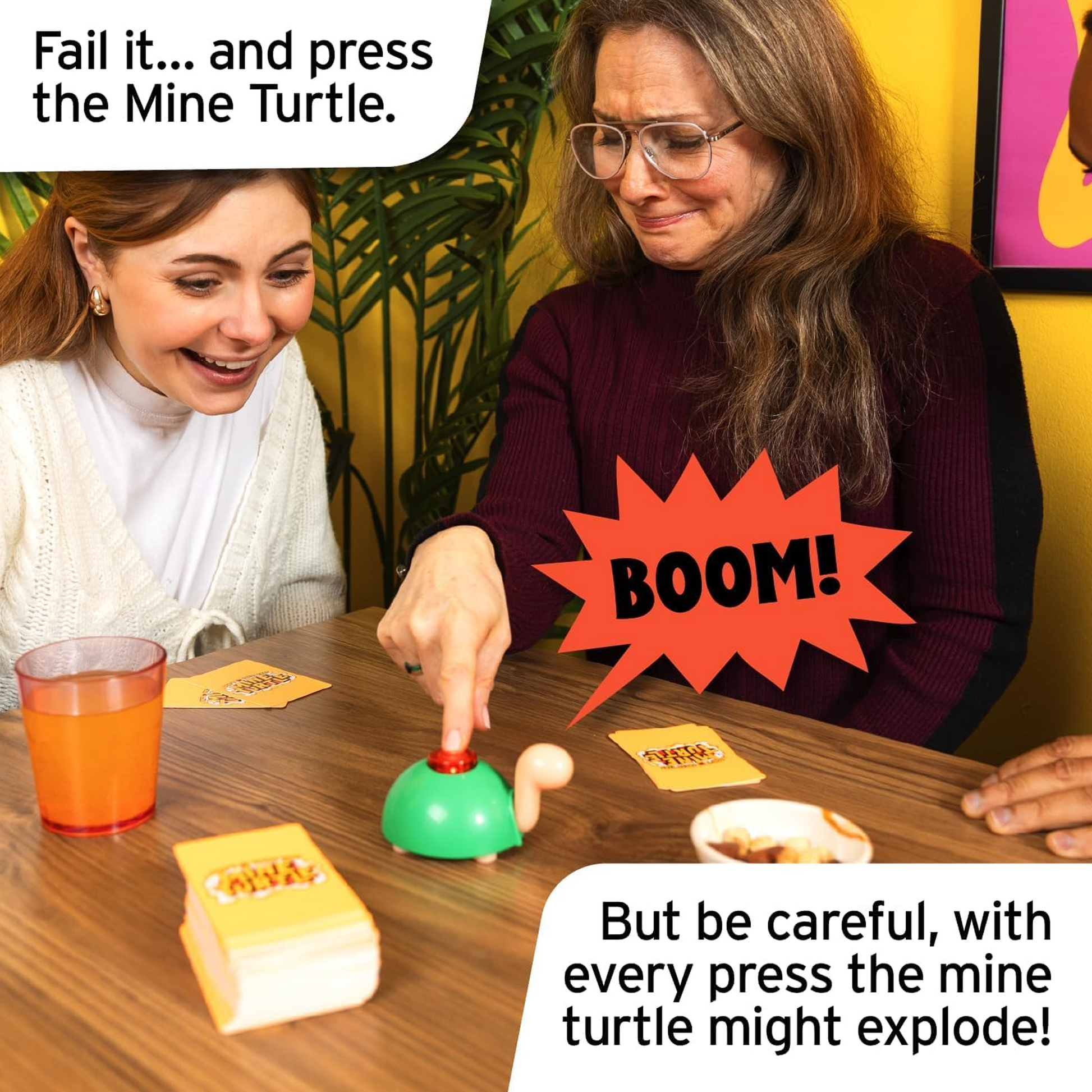 2 Women Laughing and Playing Don't Press That Mine Turtle on a Wooden Table with a Plant and Yellow Wall in the Background  | Happy Piranha