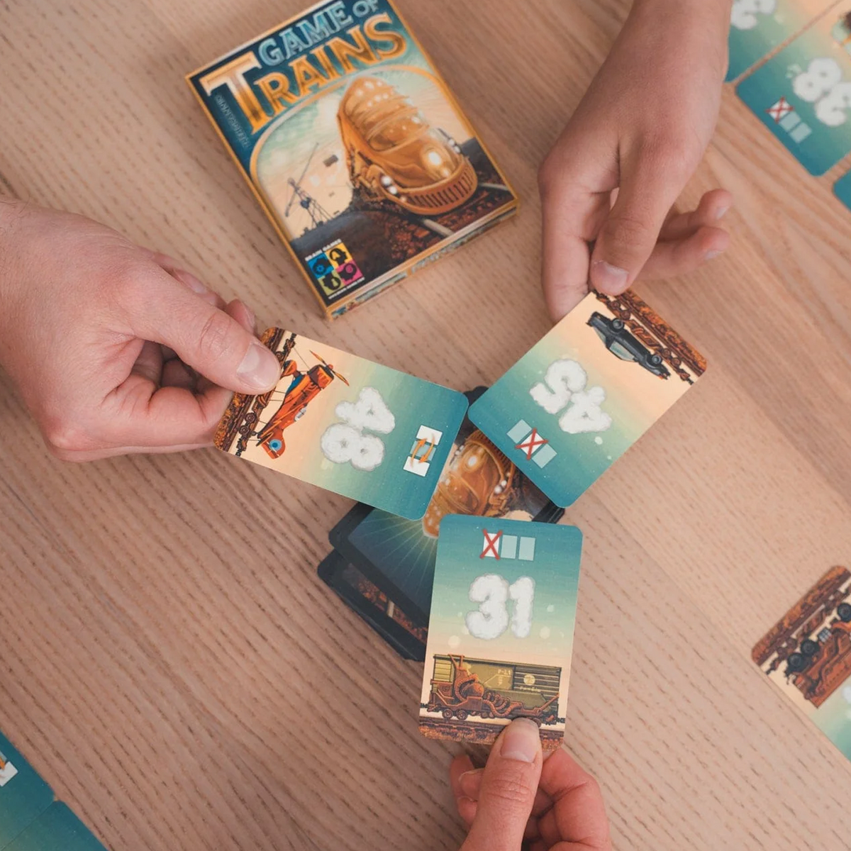 Game of Trains Card Game (A Card Being Held in the Hand of 3 Different People) on a Pine Wood Coloured Background | Happy Piranha