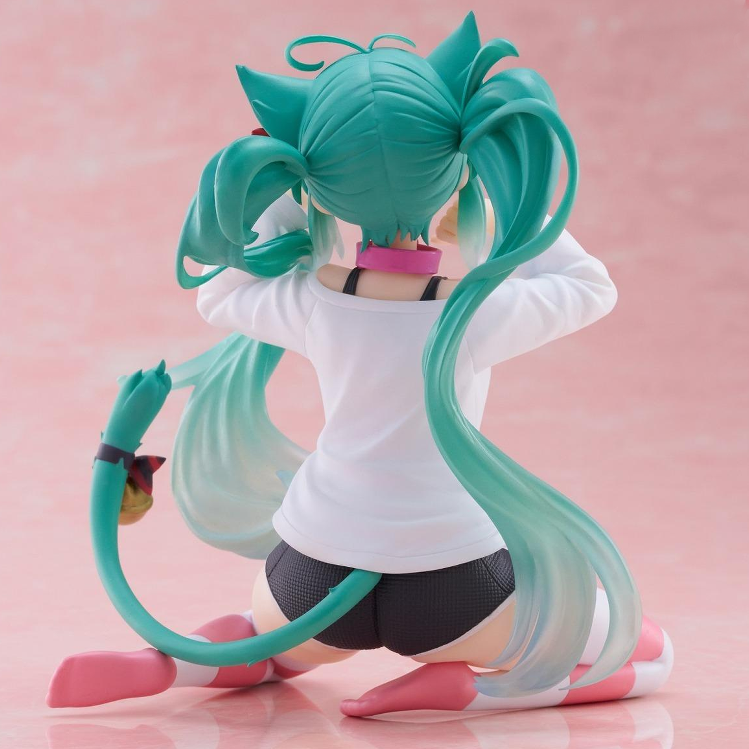 A turquoise-haired anime-style figurine shown from the back, kneeling on a pink surface against a pink background. The figure wears a white top, dark gray shorts, and pink-and-white shoes, with a long curved tail and cat-ear hair details | Hatsune Miku Desktop Cute Nekomimi T-Shirt ver. Figure | Happy Piranha