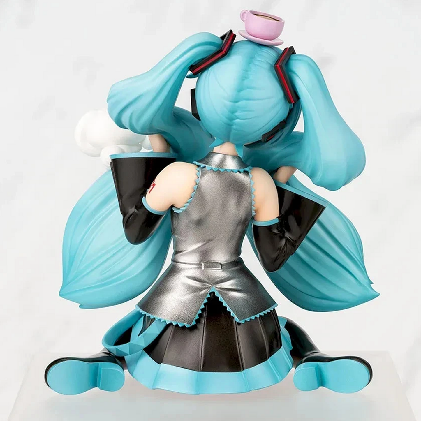 A collectible figurine of Hatsune Miku shown from the back, kneeling in a seated pose. She has signature turquoise blue pigtails with black ribbons and a pink teacup accessory on top, wearing a metallic silver and black dress with turquoise trim | Hatsune Miku x Cinnamoroll Paprio Figure Mofutto ver. | Happy Piranha