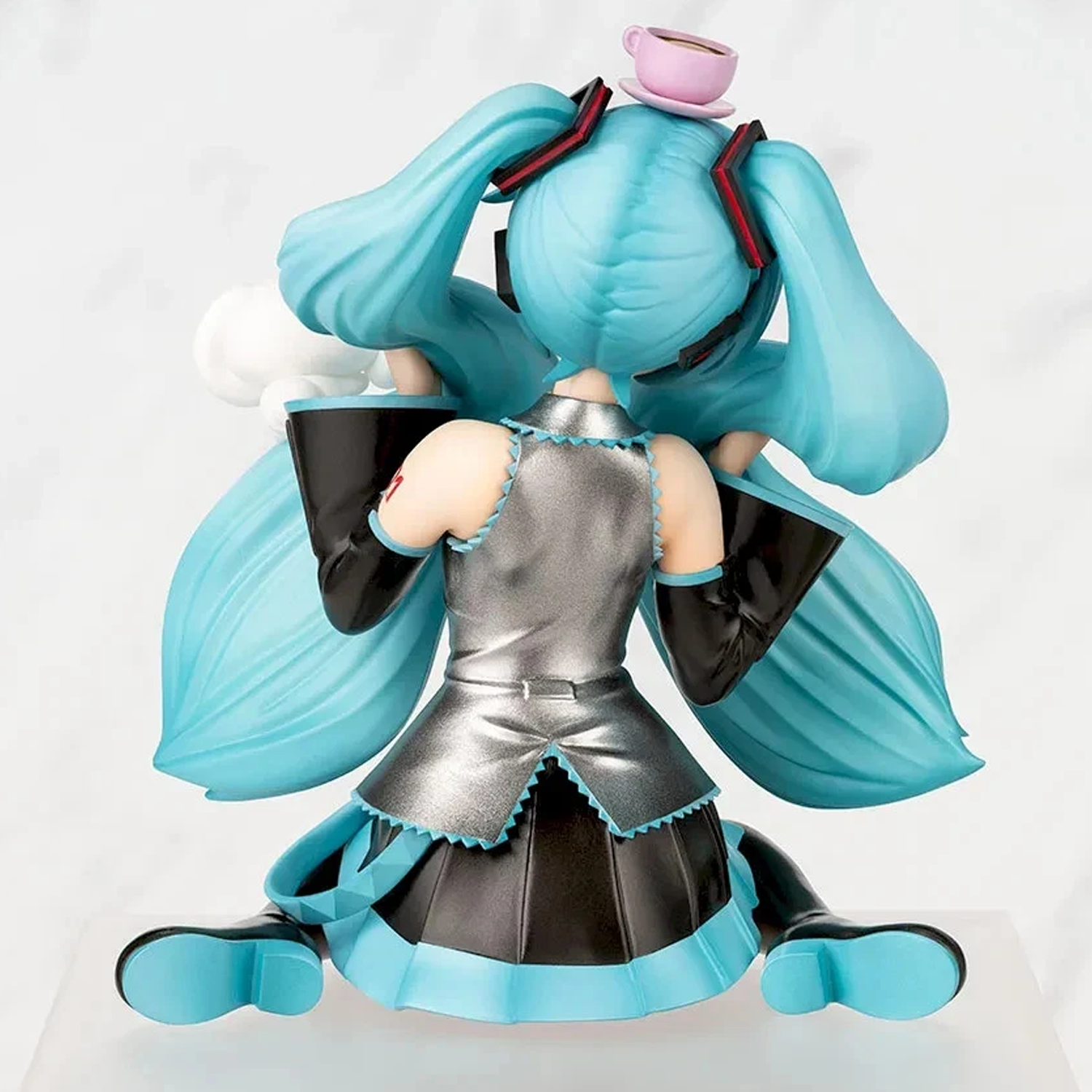 A collectible figurine of Hatsune Miku shown from the back, kneeling in a seated pose. She has signature turquoise blue pigtails with black ribbons and a pink teacup accessory on top, wearing a metallic silver and black dress with turquoise trim | Hatsune Miku x Cinnamoroll Paprio Figure Mofutto ver. | Happy Piranha