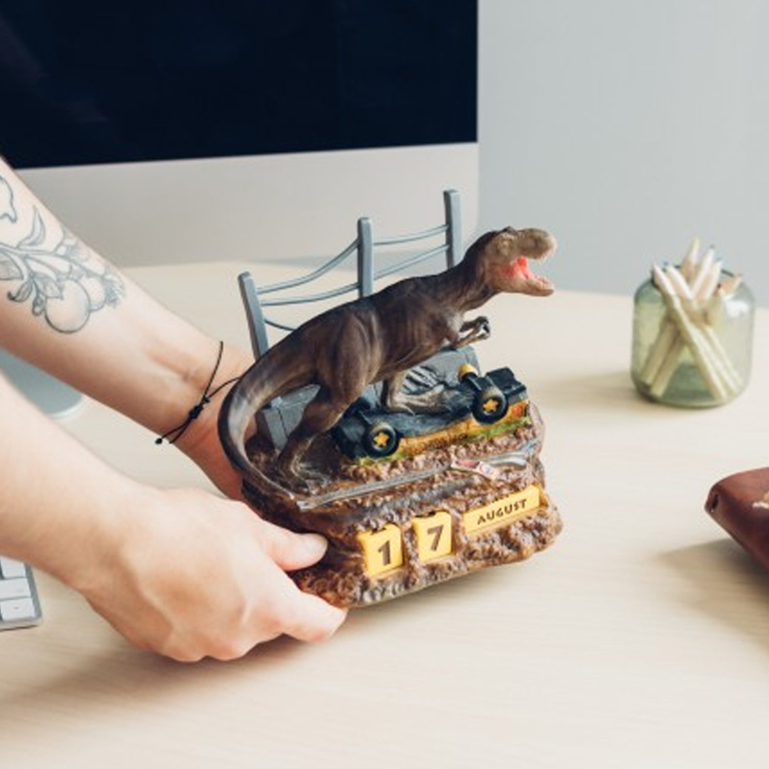 Jurassic Park T-Rex Perpetual Calendar (Front) Being Placed on a White Computer Desk by a Person with a Tattooed Arm| Happy Piranha