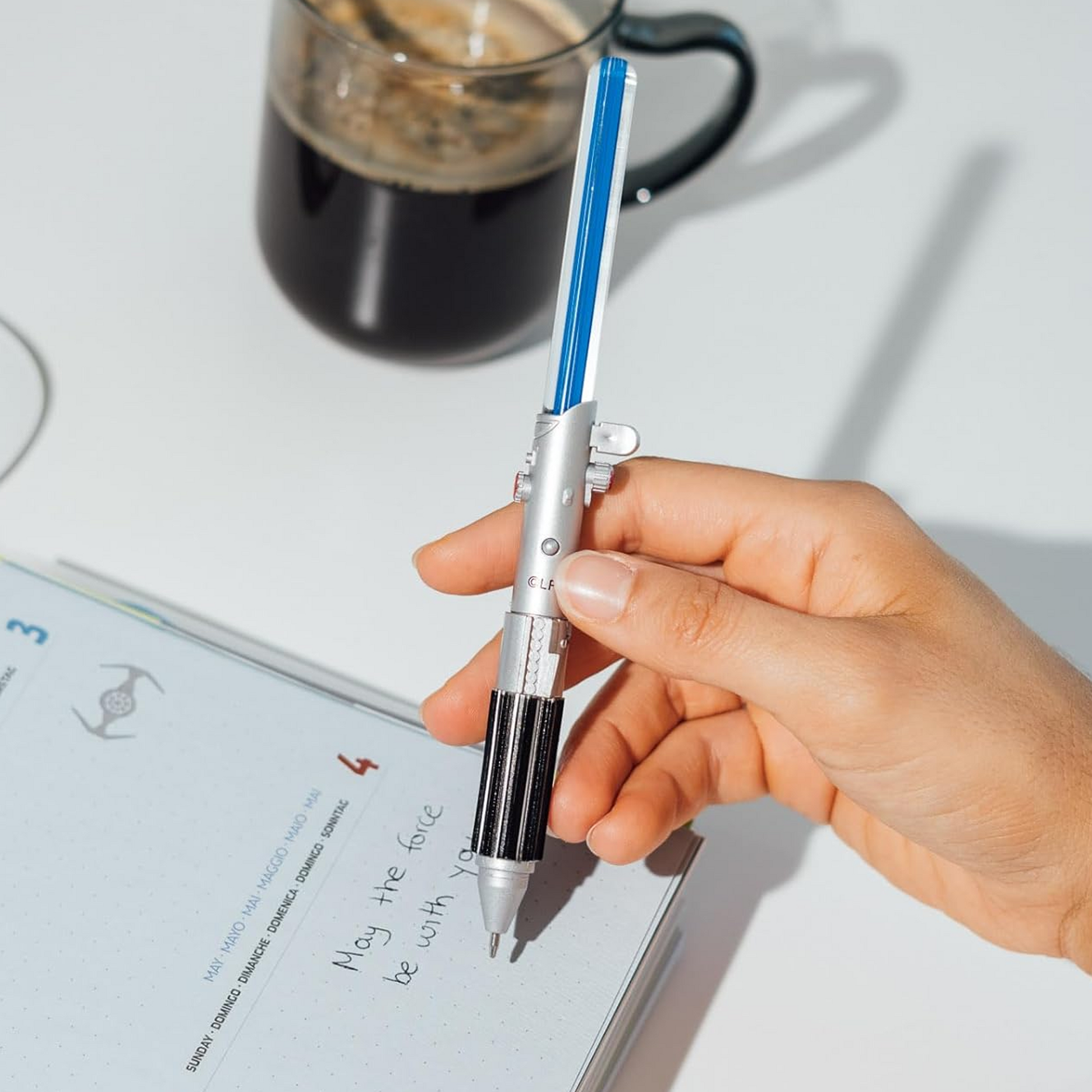 A Blue Star Wars Light Up  Lightsaber Pen in a Person's Hand Next to a Cup of Coffee on a White Table | Happy Piranha