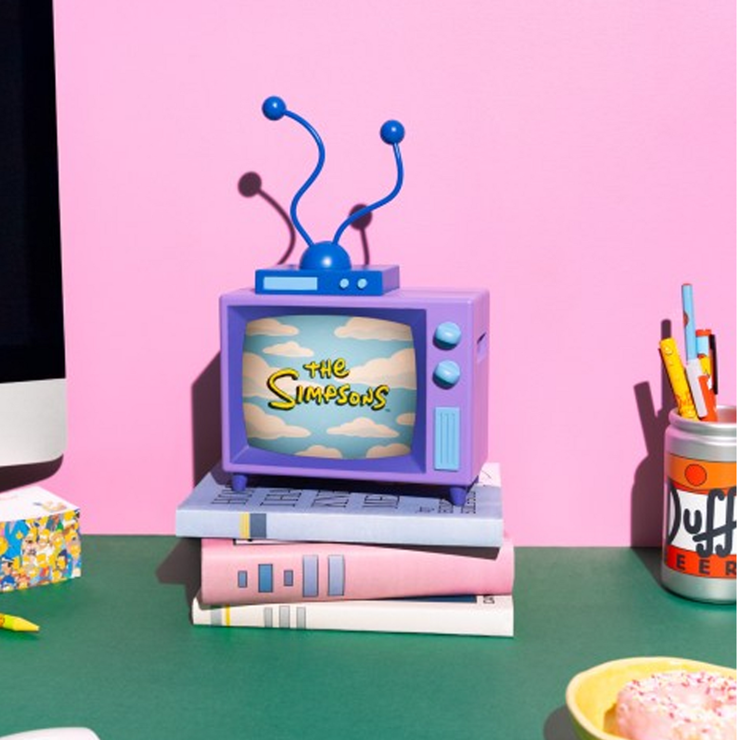 The Simpsons TV Table Lamp Turned on and Showing The Simpsons Logo, Perched on top of Some Pink and Blue books, on a Green Desktop with a Pink Wall Behind | Happy Piranha