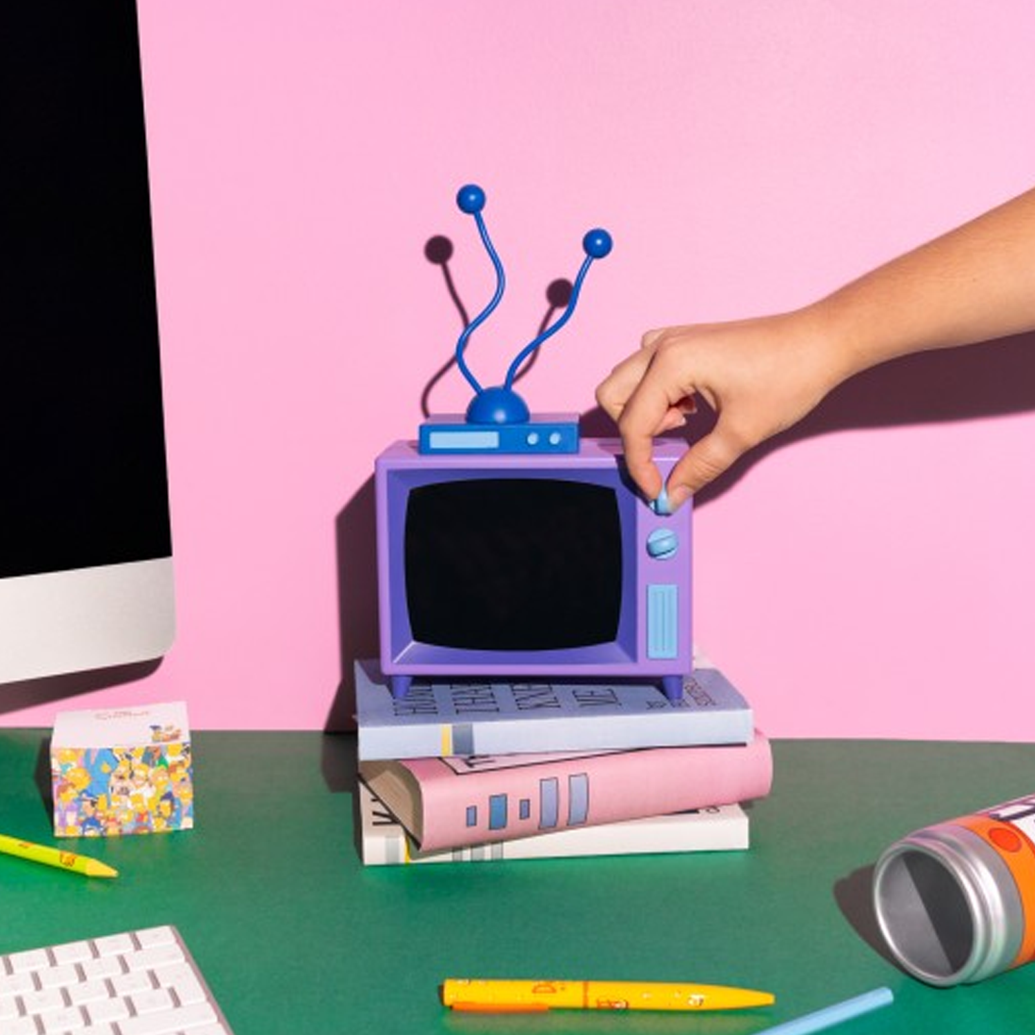 The Simpsons TV Table Lamp Being Turned on by a Person's Hand, Perched on top of Some Pink and Blue books, on a Green Desktop with a Pink Wall Behind | Happy Piranha