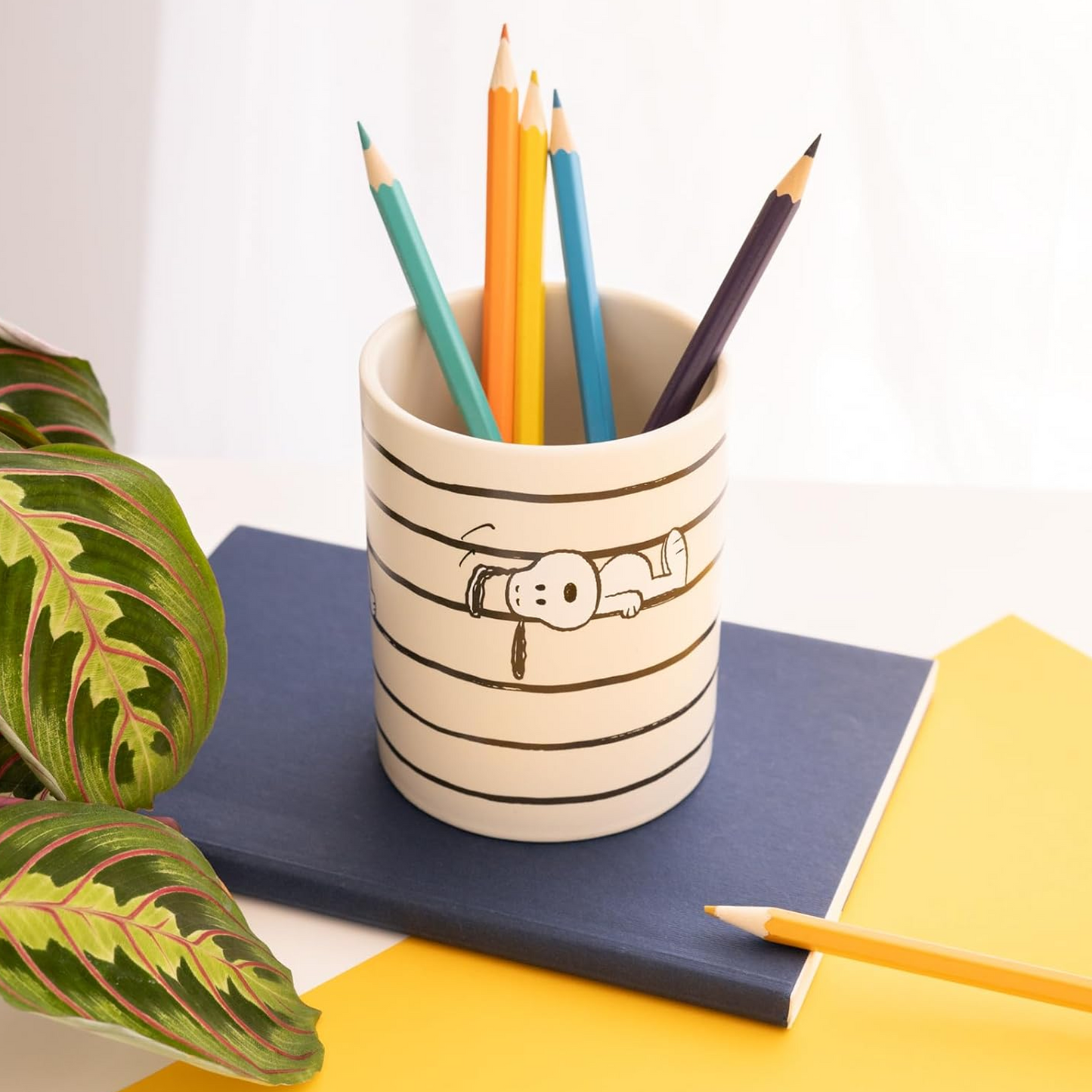Peanuts: Snoopy & Woodstock Design Pen Holder / Stationary Pot With Some Pencils Inside, on top of a Notebook on a Yellow Desk with a Green Plant in the Corner. | Happy Piranha